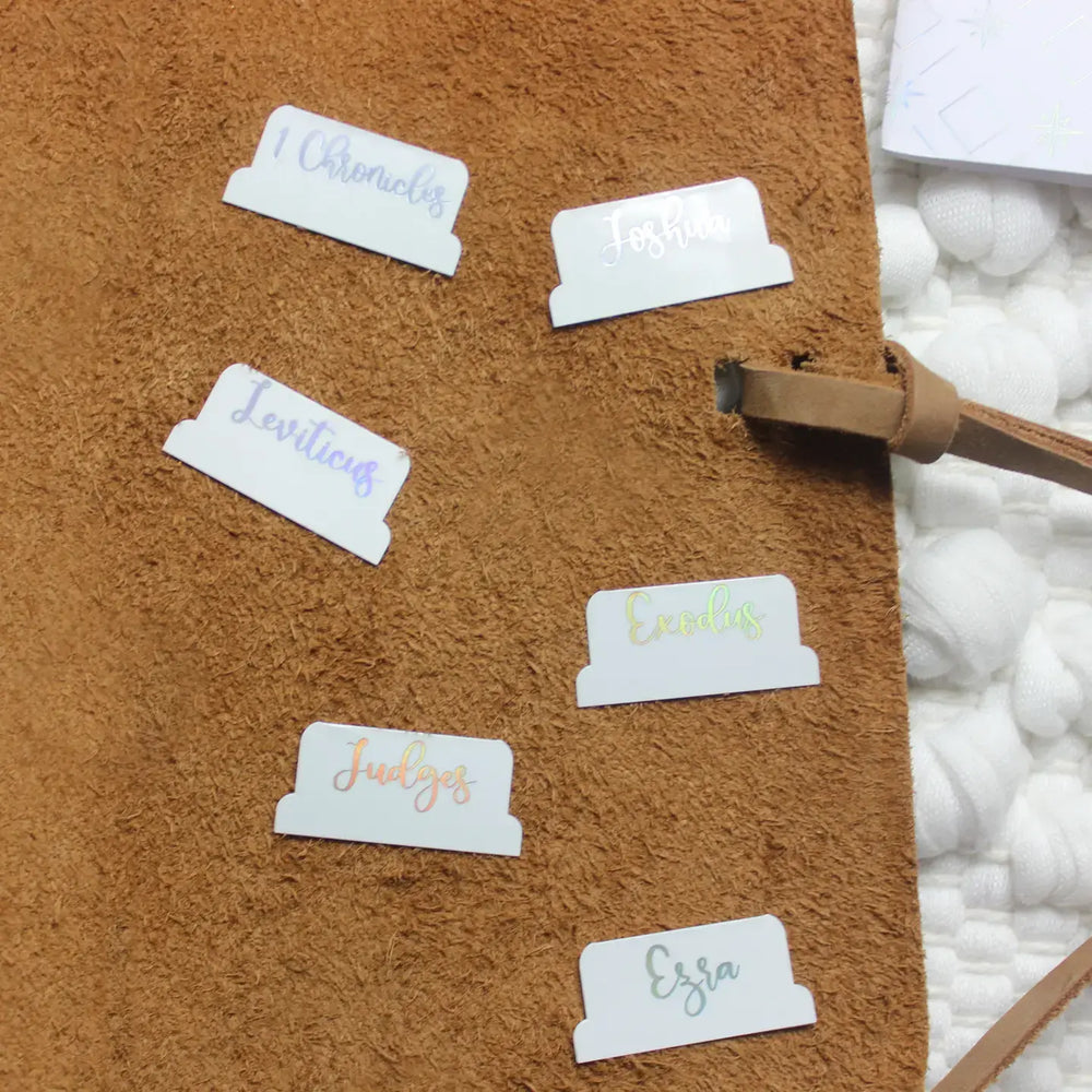 Close-up of stylish holographic Bible tabs on a brown surface, showcasing book titles for easy navigation.
