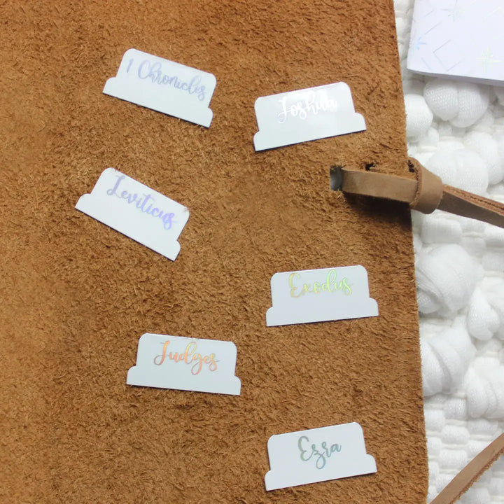 Close-up of stylish holographic Bible tabs on a brown surface, showcasing book titles for easy navigation.