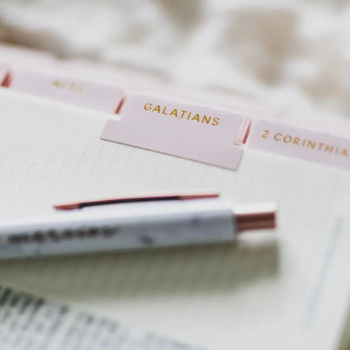 Blush Bible tabs labeled 'Galatians' and '2 Corinthians' on a notebook page with a pen, elegantly organizing scripture.