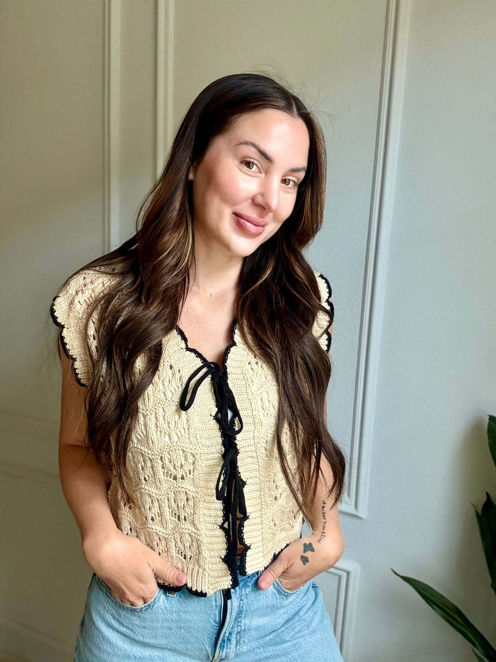 Woman wearing Faithful Threads Crochet Sweater Vest with bow tie detail, showcasing cozy elegance in a bright setting.