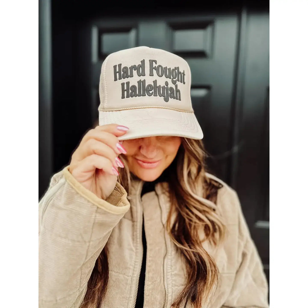 Tan trucker hat with 'Hard Fought Hallelujah' text, worn by model, celebrating resilience and faith.