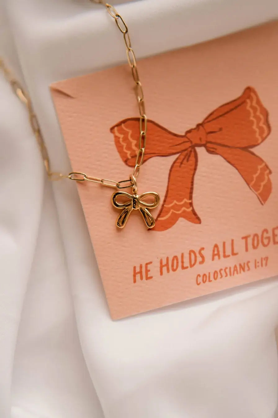 Gold bow charm necklace with a card reading 'He Holds All Together - Colossians 1:17', symbolizing faith and comfort.