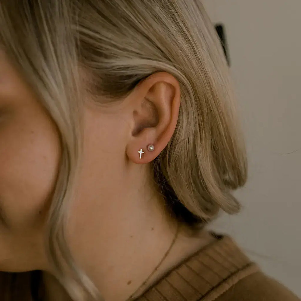 Close-up of a woman wearing Known Stud Earrings, featuring a gold cross design and a pearl, symbolizing unconditional love.