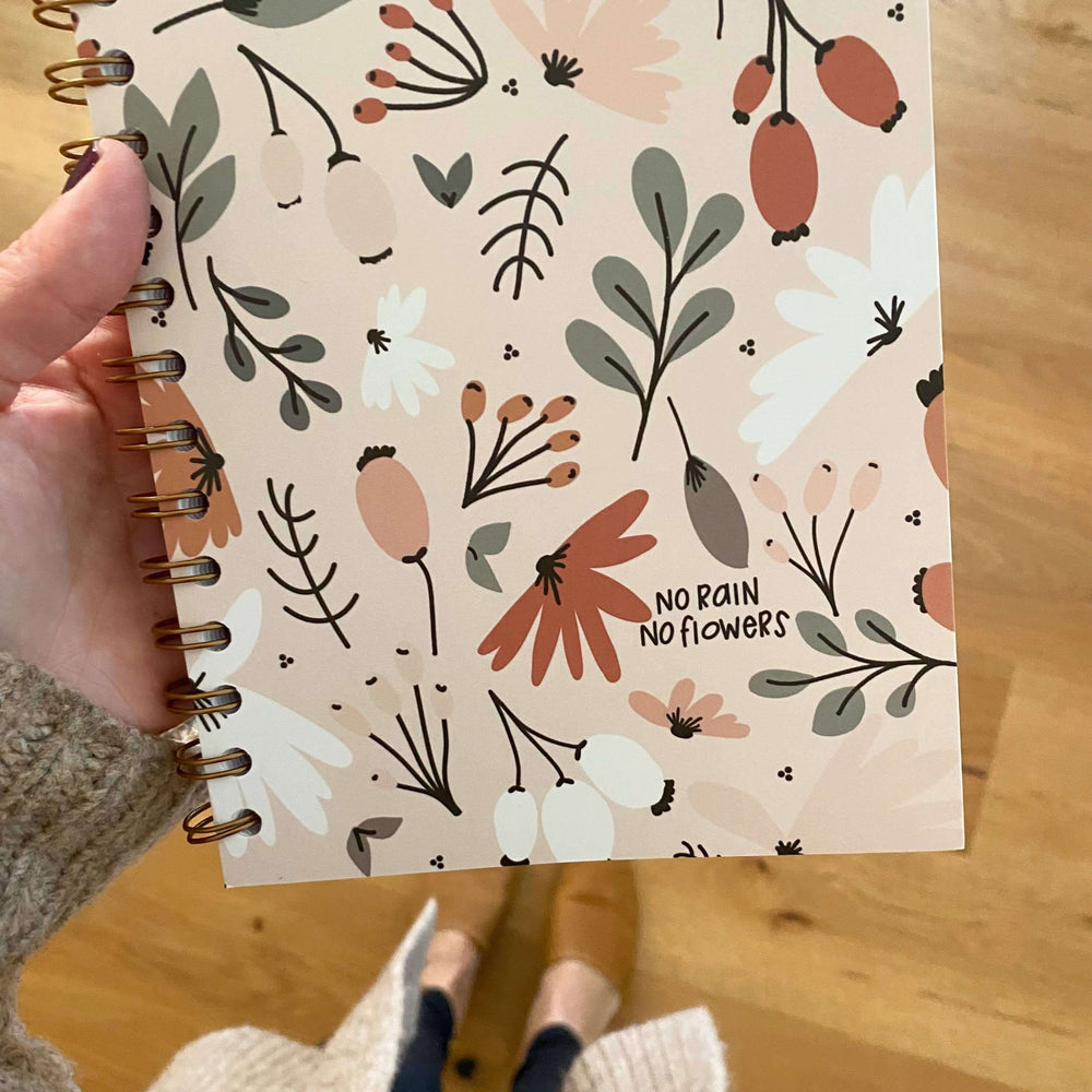 Hand holding a spiral-bound notebook featuring a floral design and the quote 'No Rain No Flowers'.