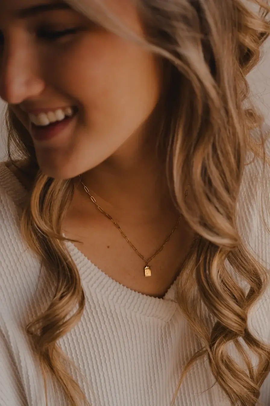 Young woman wearing gold With You Mini Tag Necklace, smiling, with soft, wavy hair.